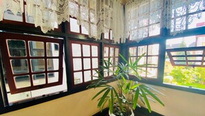 Ein Zimmer mit einem Blumentopf auf einem Tisch, Spitze an den Fenstern und Blick ins Grüne.