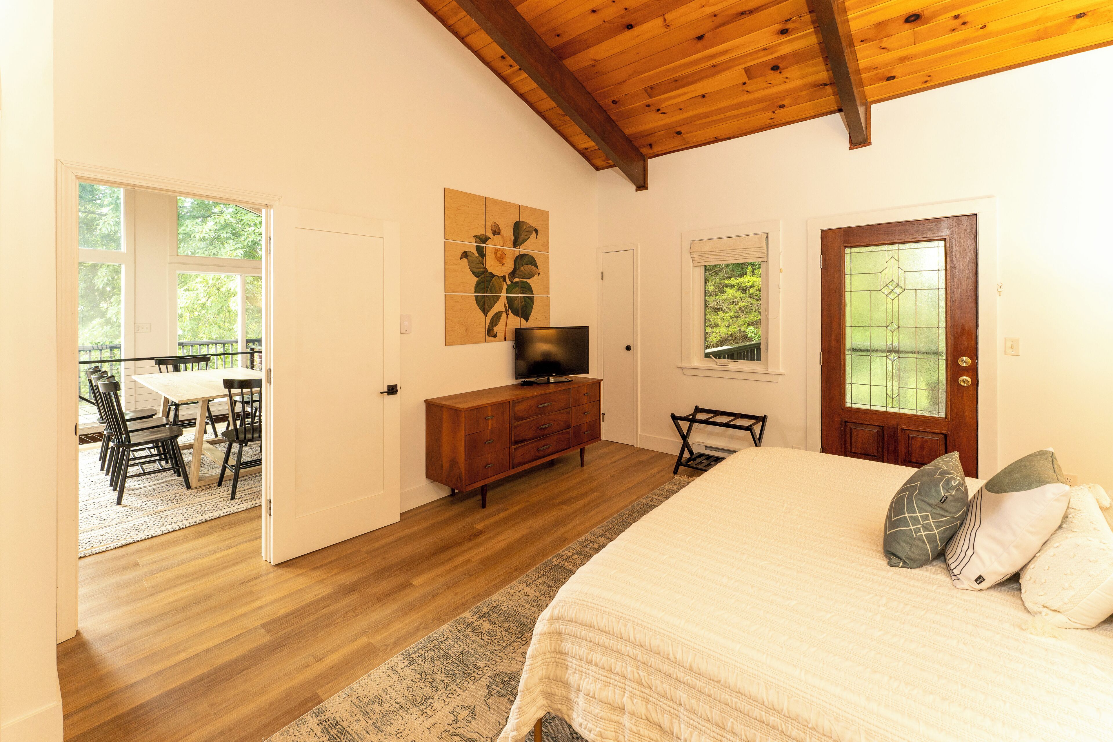 A bedroom with a wooden ceiling, a bed with pillows, a TV on a wooden dresser, and a dining area with chairs and a table.