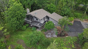 A two-story house with a deck, surrounded by dense greenery. - Enjoy Cabin Vibes & Lake Views, Close to Town, Lake & FLX Activities; EV Charger (Canandaigua)