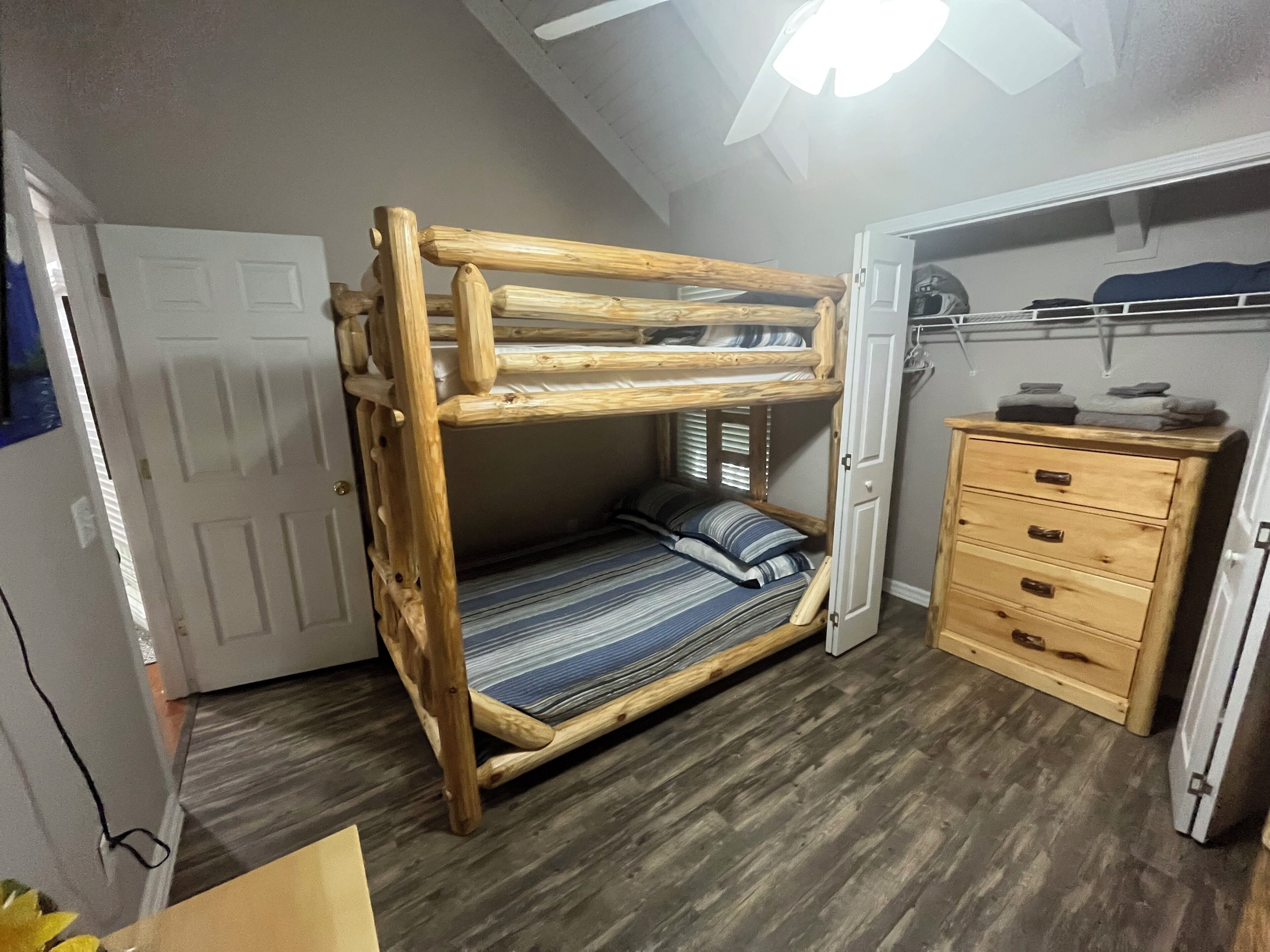 A bunk bed with a wooden frame, a dresser with drawers, and a ceiling fan in a room with wooden flooring.
