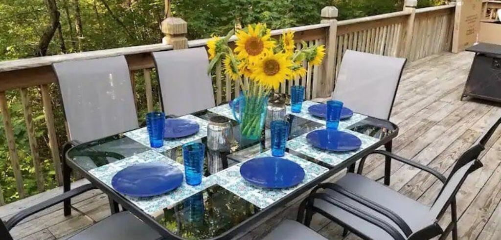 Ett bord dukat för fyra personer, med blå tallrikar och glas, en vas med solrosor och en träveranda med ett räcke.