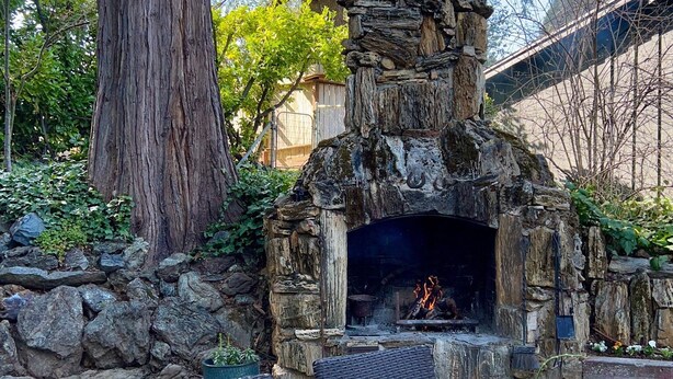 An outdoor stone fireplace with a fire, surrounded by a stone wall and a large tree.