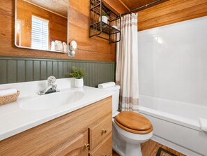 Une salle de bain avec un meuble-lavabo en bois, un lavabo blanc, des toilettes avec un abattant en bois, un rideau de douche et un miroir.