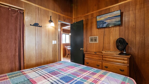 Ein Schlafzimmer mit kariertem Bettbezug, Holzwänden, einem Schminktisch, einem Ventilator und einem Bild an der Wand.