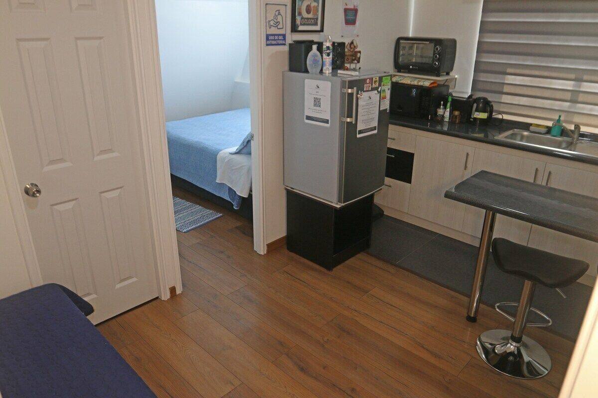 A compact kitchen with a refrigerator, microwave, and sink. There's a small bar stool and a bed with a blue blanket in the background.