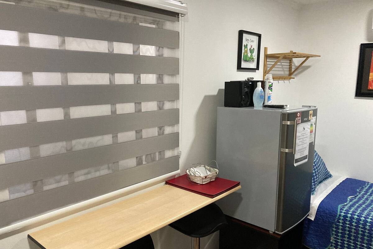 A compact room with a small desk, a refrigerator, a bed with a blue and white patterned bedspread, and a window with horizontal blinds.