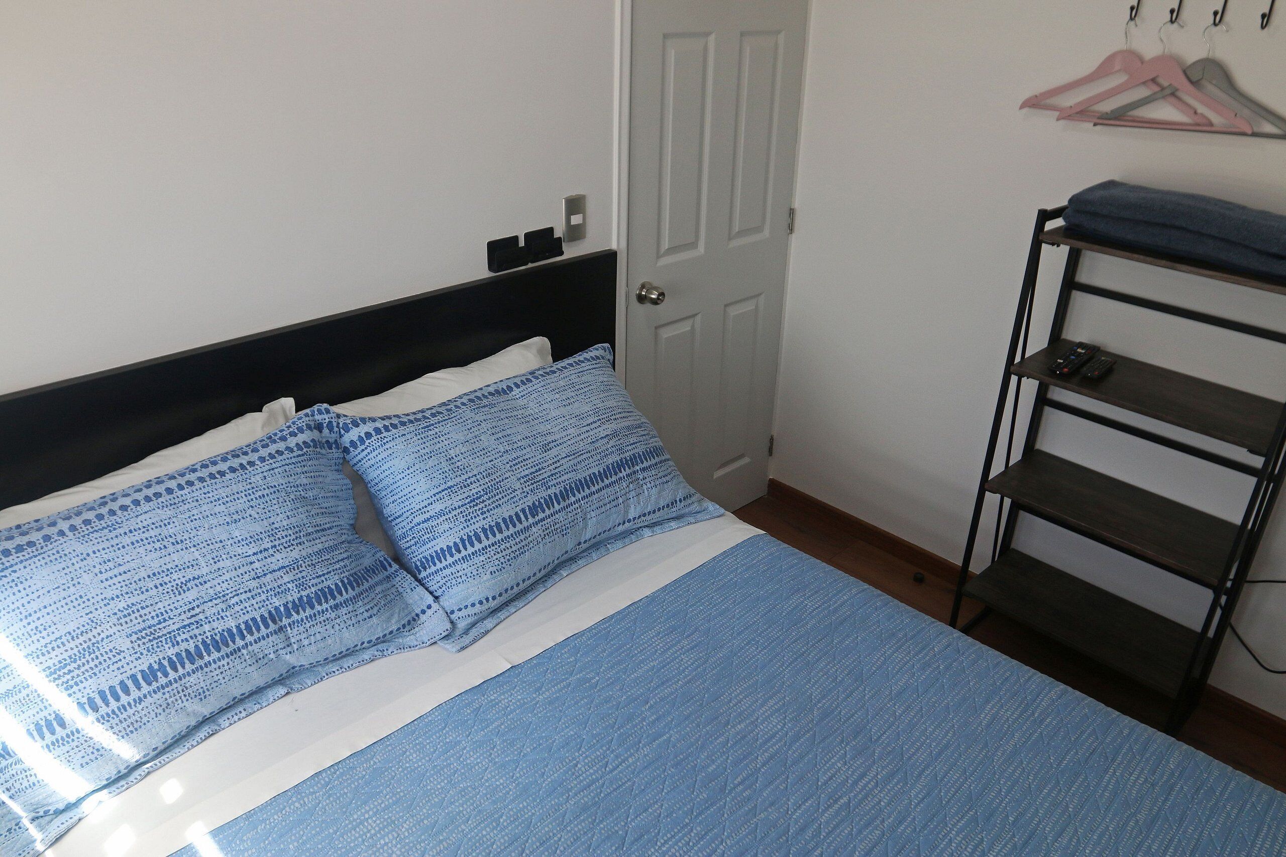 Een net opgemaakt bed met blauw beddengoed, een witte deur en een metalen plank met kledinghangers.