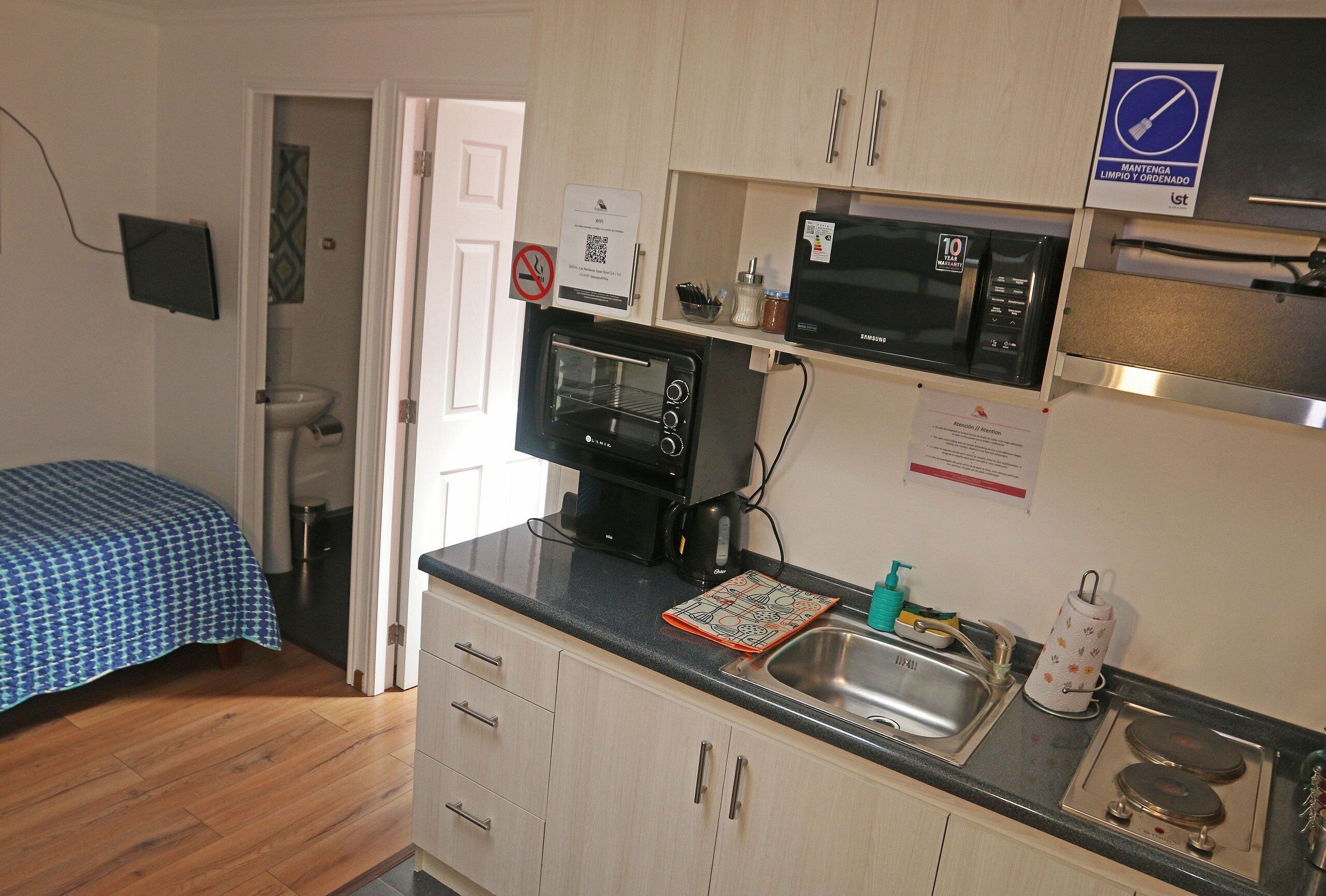Una cocina compacta con microondas, horno y lavamanos. En la habitación contigua hay una cama con colcha azul a cuadros.
