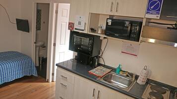 Una cocina compacta con microondas, horno y lavamanos. En la habitación contigua hay una cama con colcha azul a cuadros.