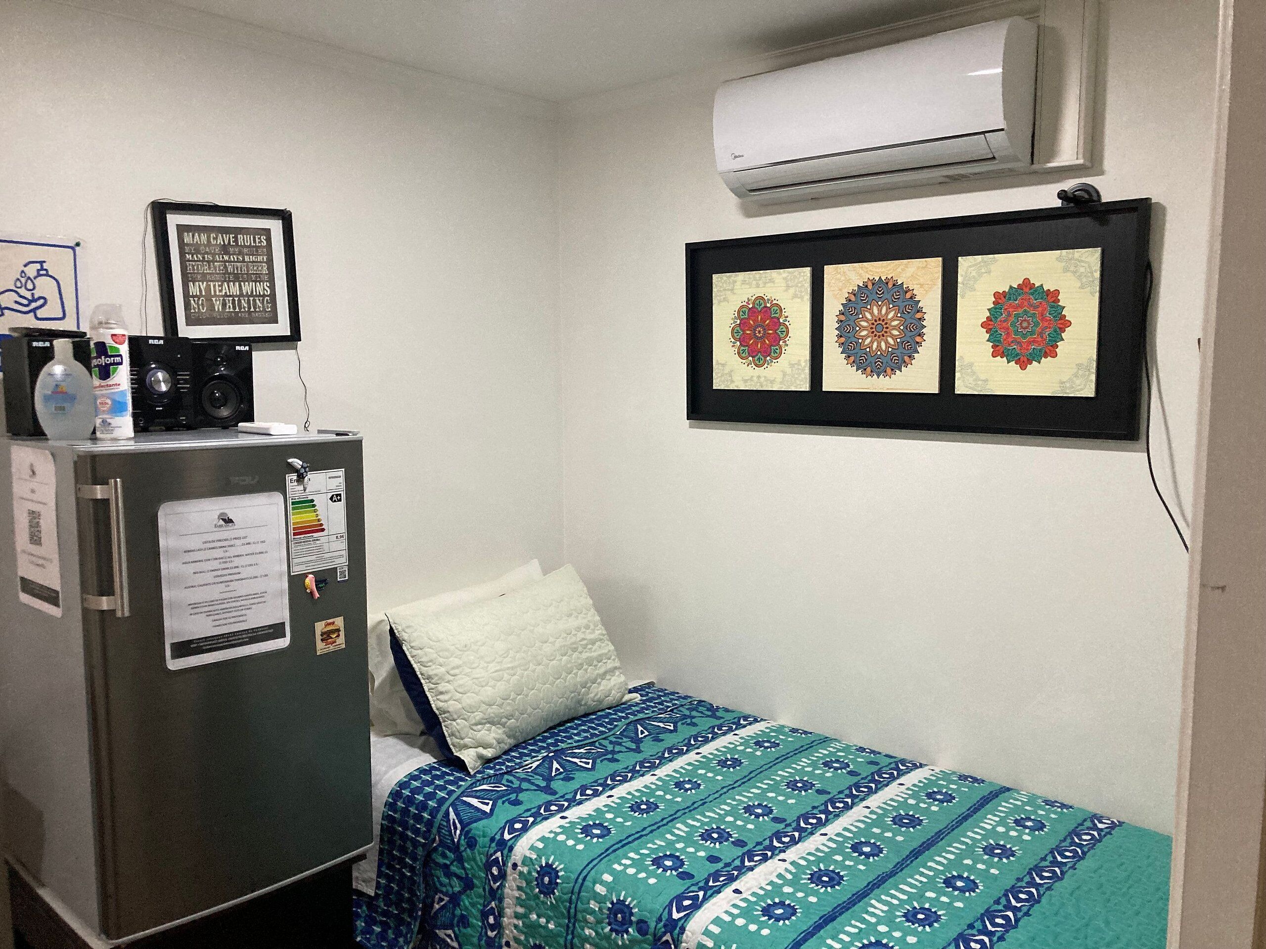 A small room with a bed, a refrigerator, and an air conditioner. There are framed pictures on the wall and a bottle of hand sanitizer on the refrigerator.