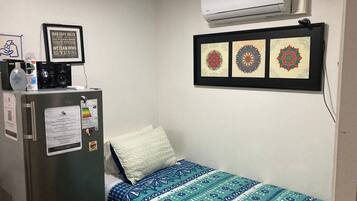 A small room with a bed, a refrigerator, and an air conditioner. There are framed pictures on the wall and a bottle of hand sanitizer on the refrigerator.