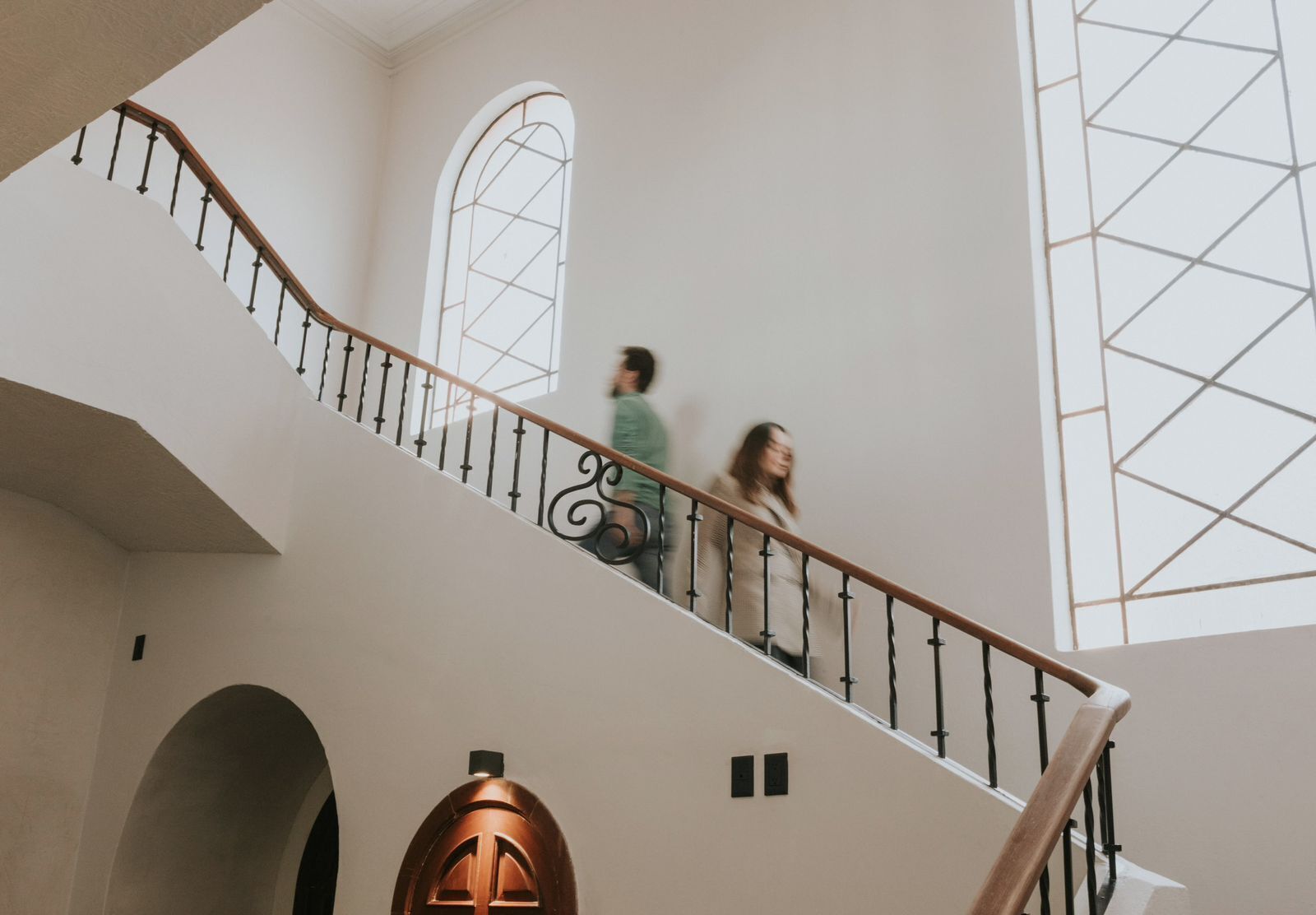 A staircase with a wooden handrail and black balusters, leading to a landing with a large arched window and a geometric patterned wall.