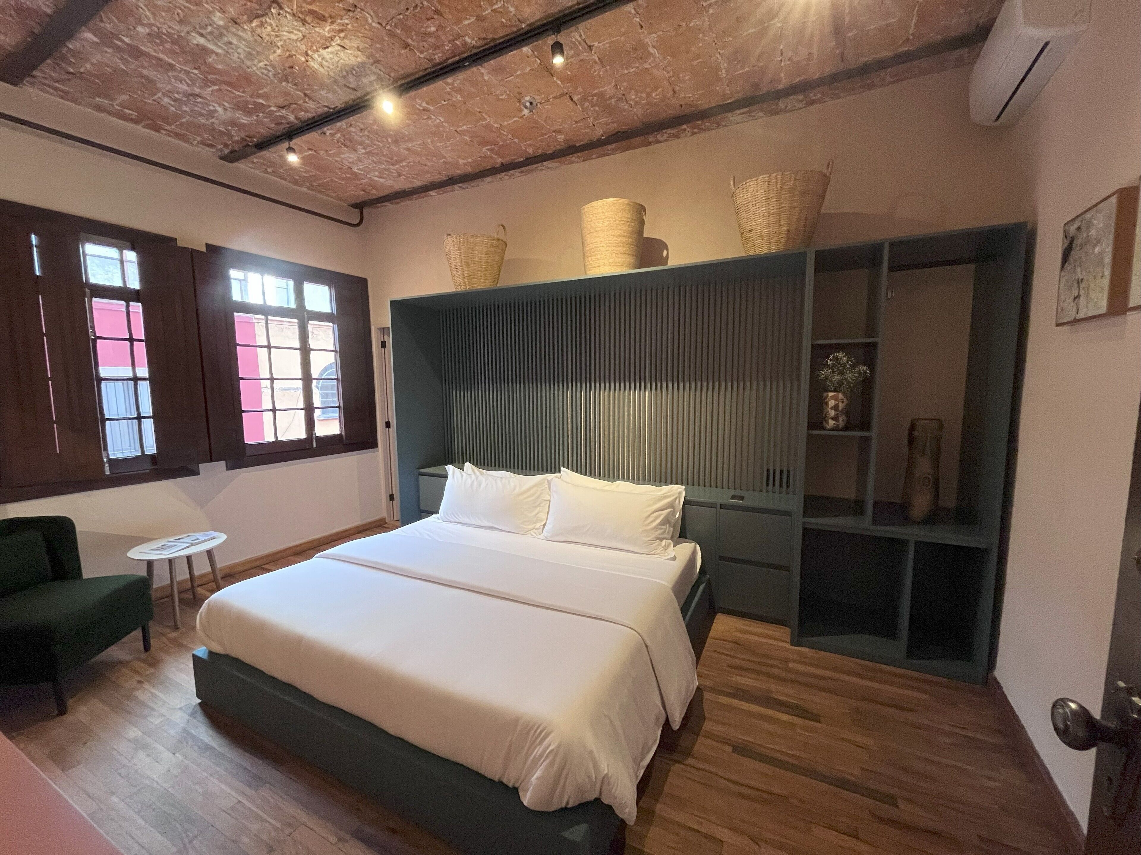A bedroom with a large bed, a green armchair, a small table, and a wardrobe with baskets on top.