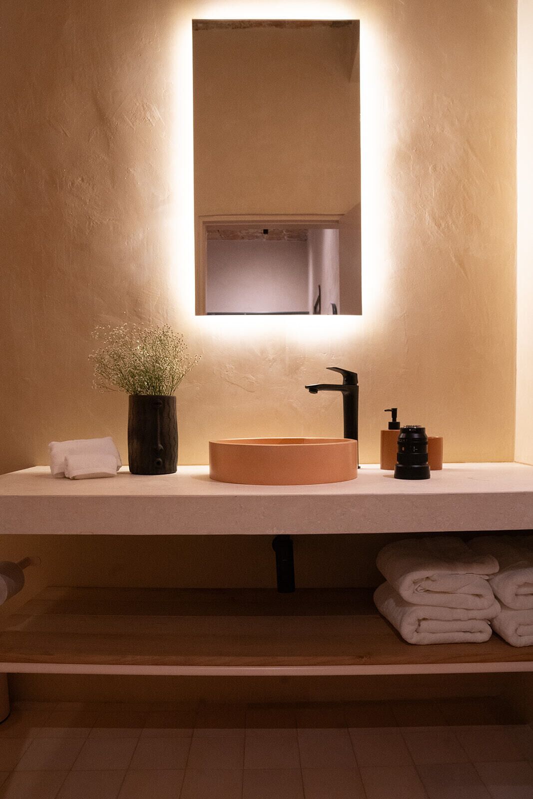 Un baño con un lavamanos redondo, un espejo, una planta en maceta, un dispensador de toallas y una estantería con toallas dobladas.