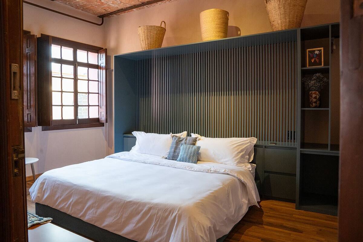 Ein ordentlich bezogenes Bett mit weißen Leinen, ein Holzboden, ein Fenster mit Jalousien und ein blaues Kopfende mit vertikalen Lamellen.