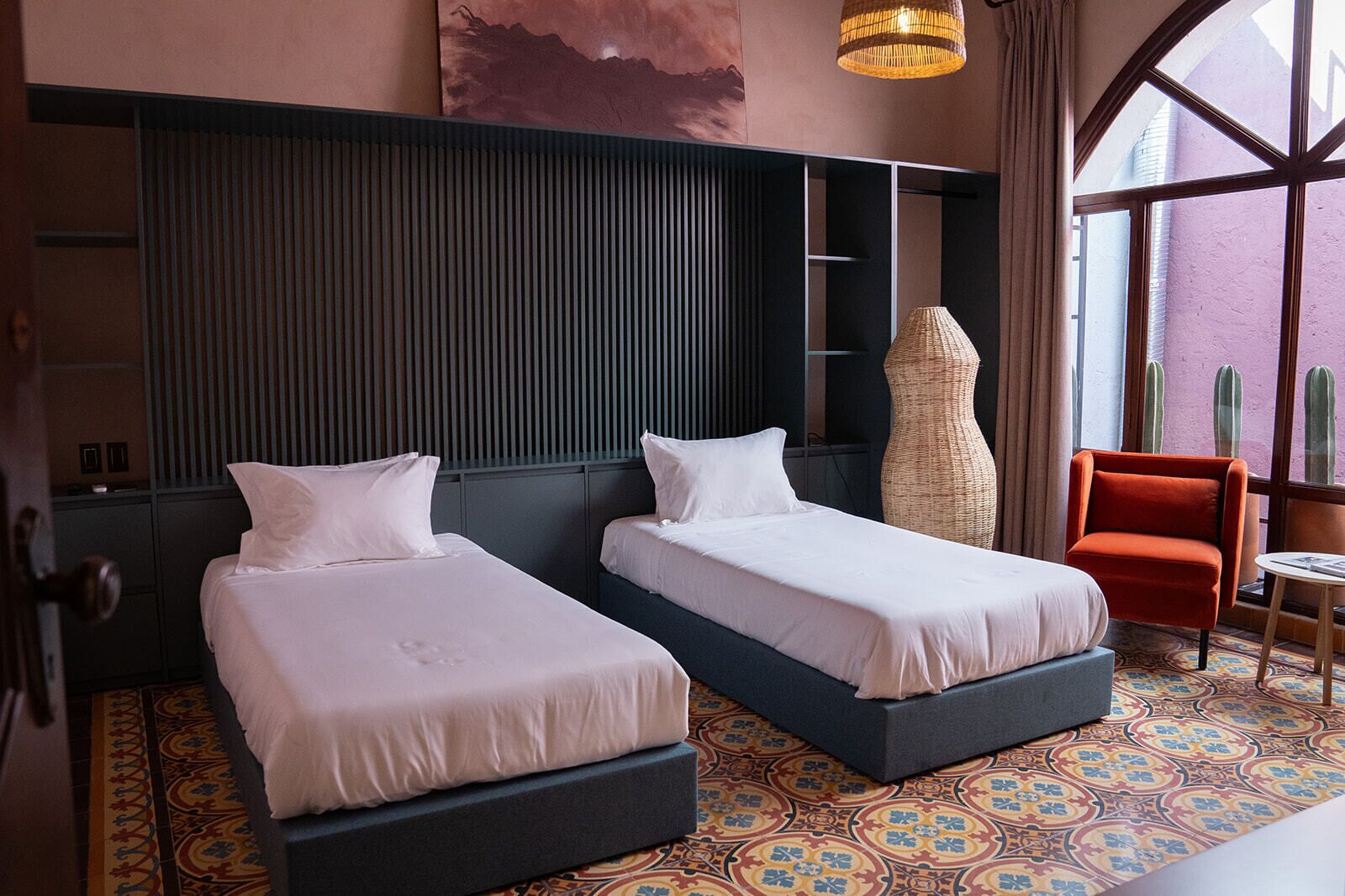 A hotel room with two beds, a large window with a view of cacti, a wicker chair, and a decorative wall piece.