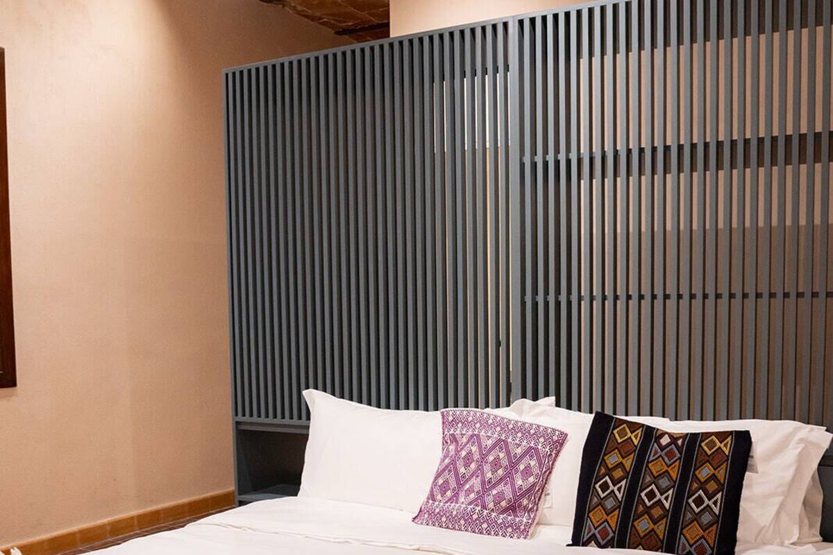Ein ordentlich bezogenes Bett mit einer gemusterten Tagesdecke, ein Kopfteil mit vertikalen Lamellen und eine schlichte Wand.