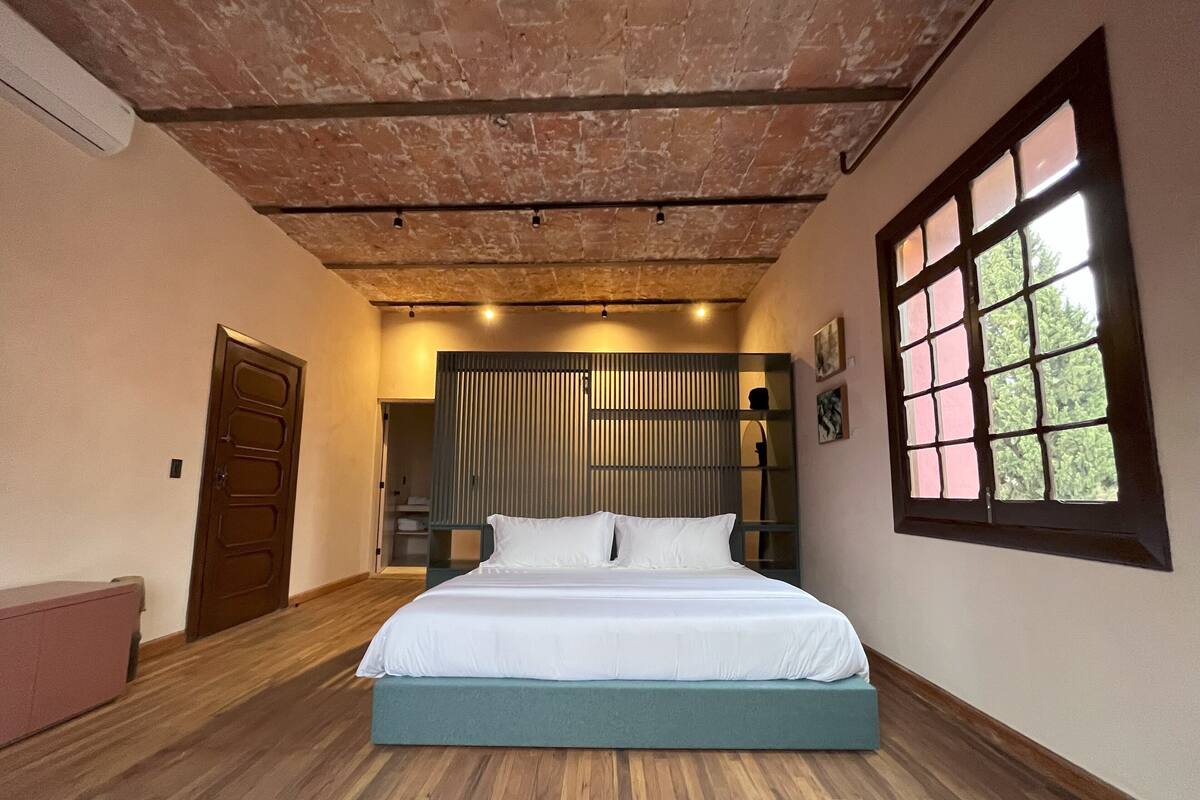 Ein Schlafzimmer mit Holzboden, einem Bett mit weißer Bettwäsche, einem großen Fenster mit Blick auf Bäume und einer Ziegeldecke.