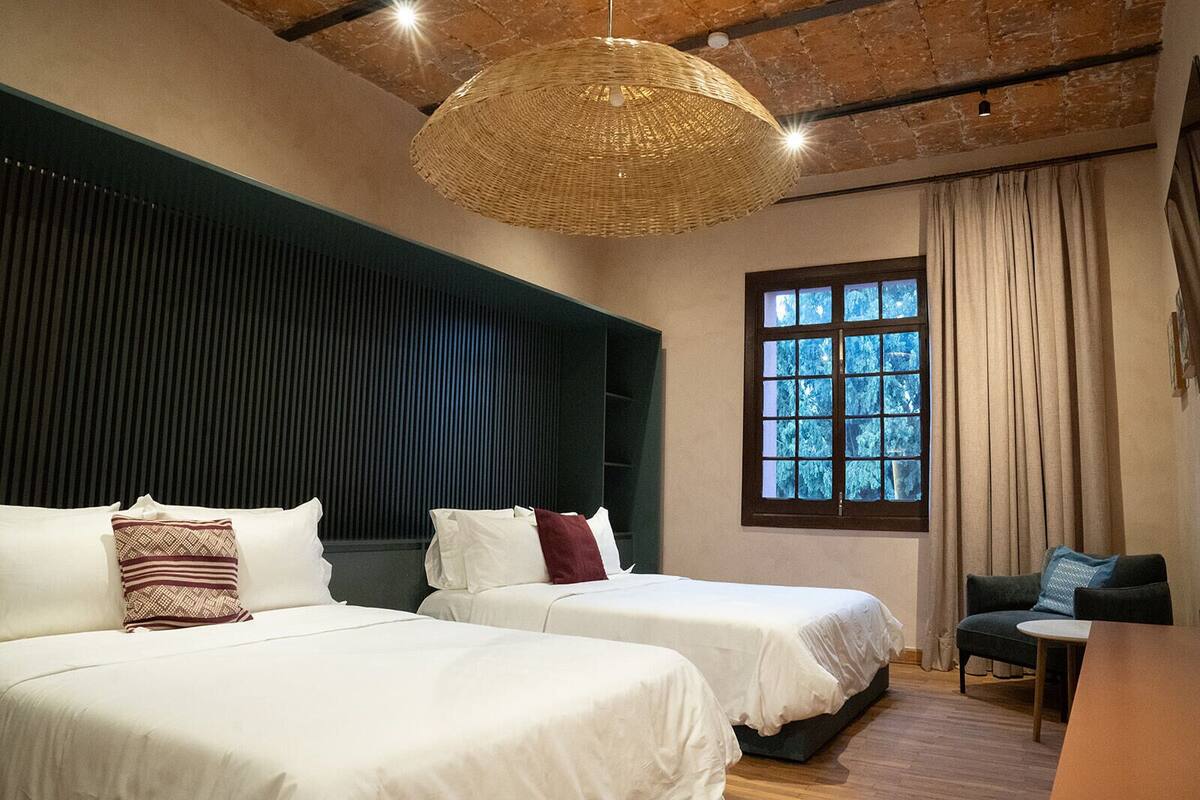 Ein Hotelzimmer mit zwei Betten, einer großen gewebten Leuchte, einem Fenster mit Vorhängen und einem kleinen Tisch.