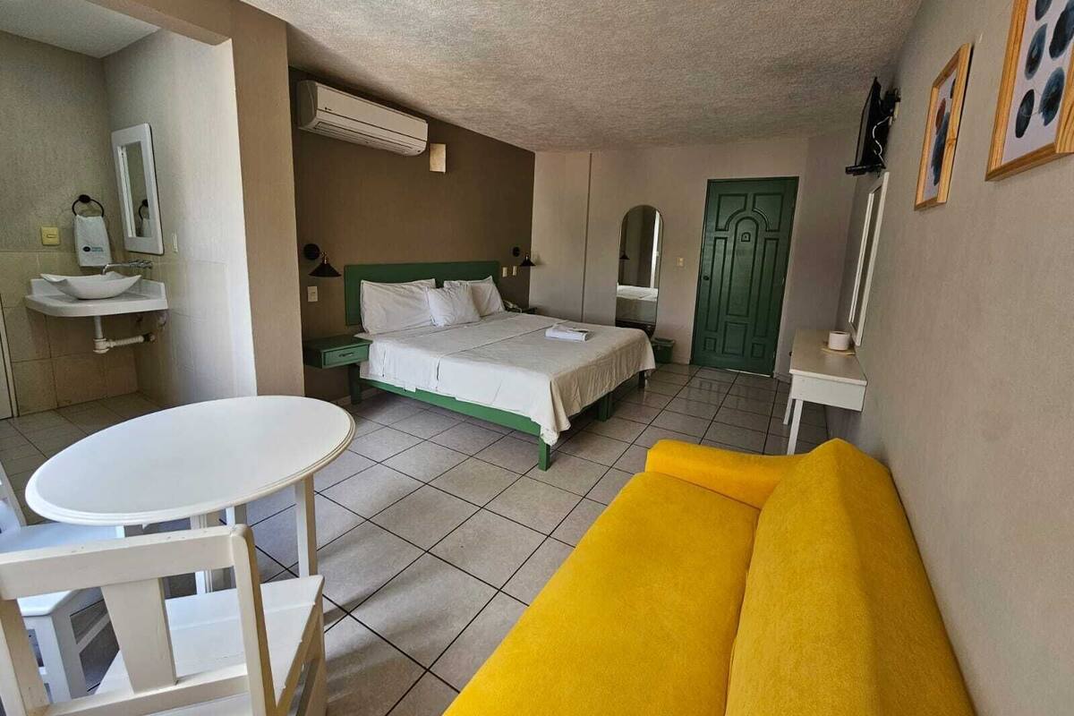 A hotel room with a bed, a round table, a yellow sofa, a sink, and a mirror.