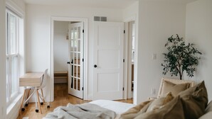 Ein Schlafzimmer mit einem Holztisch, einem Bett mit beigefarbener Bettwäsche, einer Topfpflanze und einer weißen Tür.