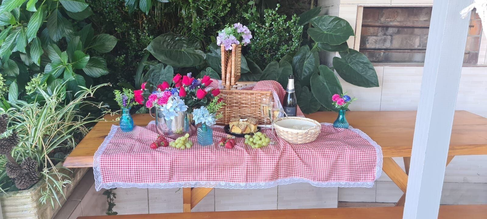 A table set outdoors with a checkered tablecloth, various fruits, flowers, and a bottle.