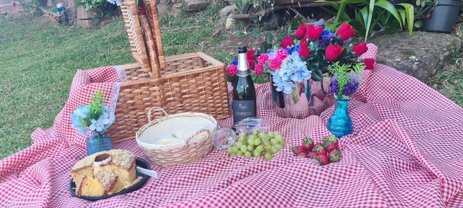 Een picknickopstelling met een rieten mand, een fles wijn, een mand met brood, een schaal druiven en een boeket bloemen op een geruit kleed.