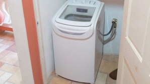A compact, white washing machine in a small, tiled room with a door on the right and a shelf above it.