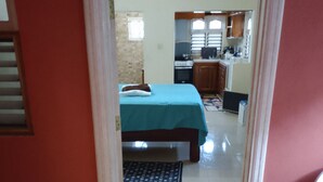 A bedroom with a bed, a bedside table, and a window. There is a kitchen area visible in the background.
