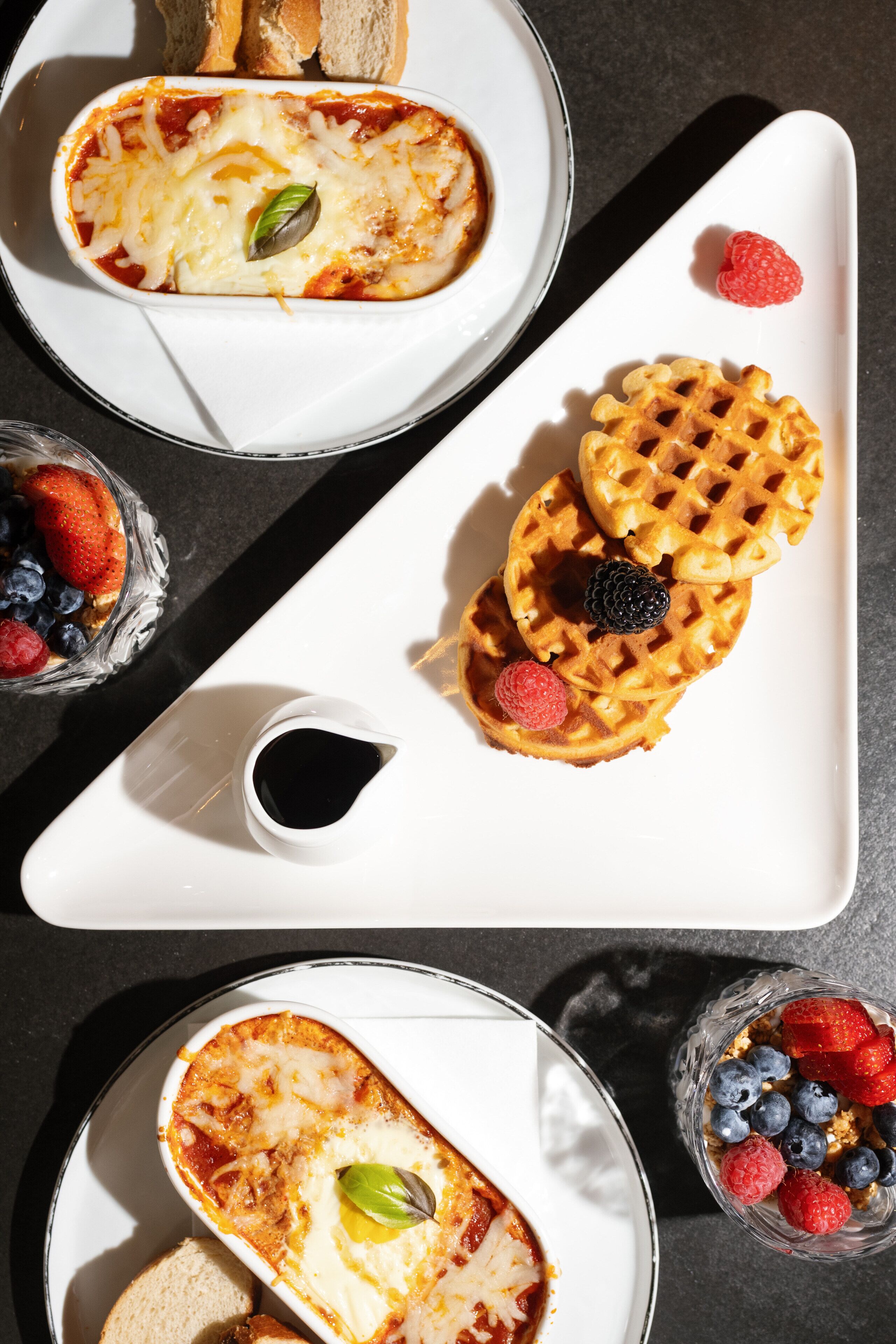 A breakfast setting with a plate of waffles topped with berries, a bowl of soup, and a small bowl of syrup.