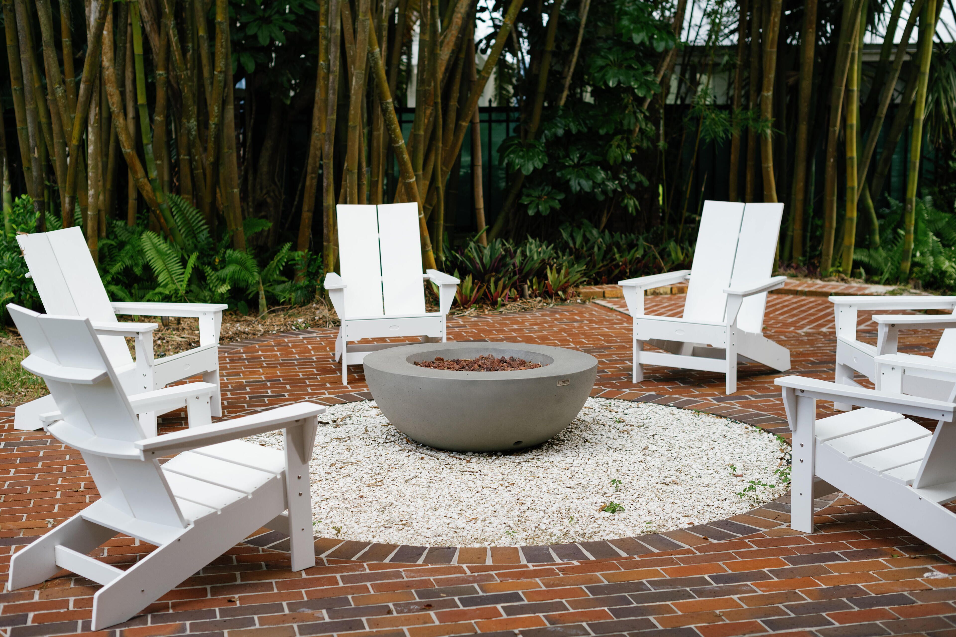 A circular fire pit surrounded by four white Adirondack chairs on a brick patio.