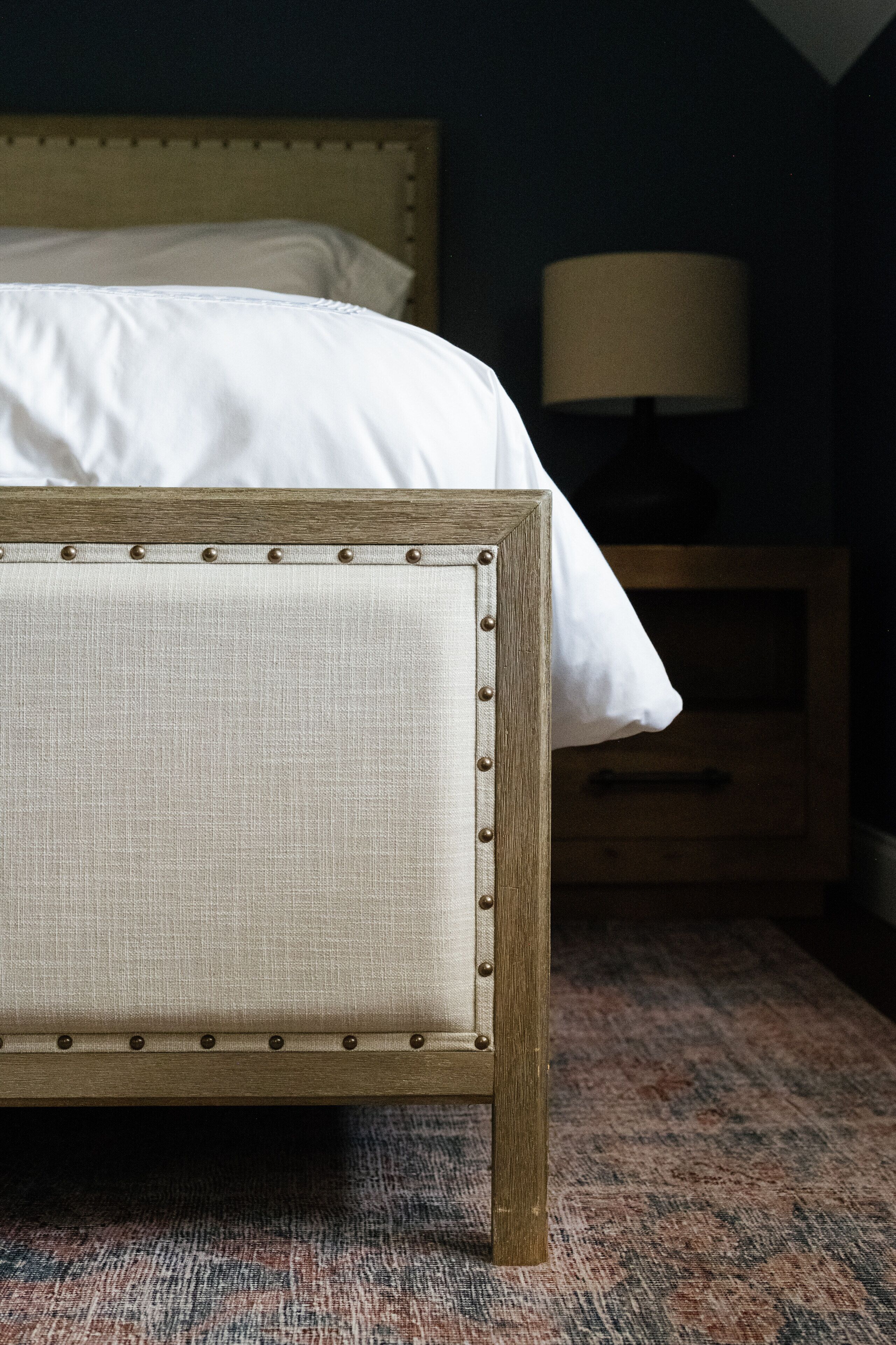 Ein Bett mit Holzgestell und Stoffpolster, ein Nachttisch aus Holz mit Lampe sowie ein Teppich mit Muster.