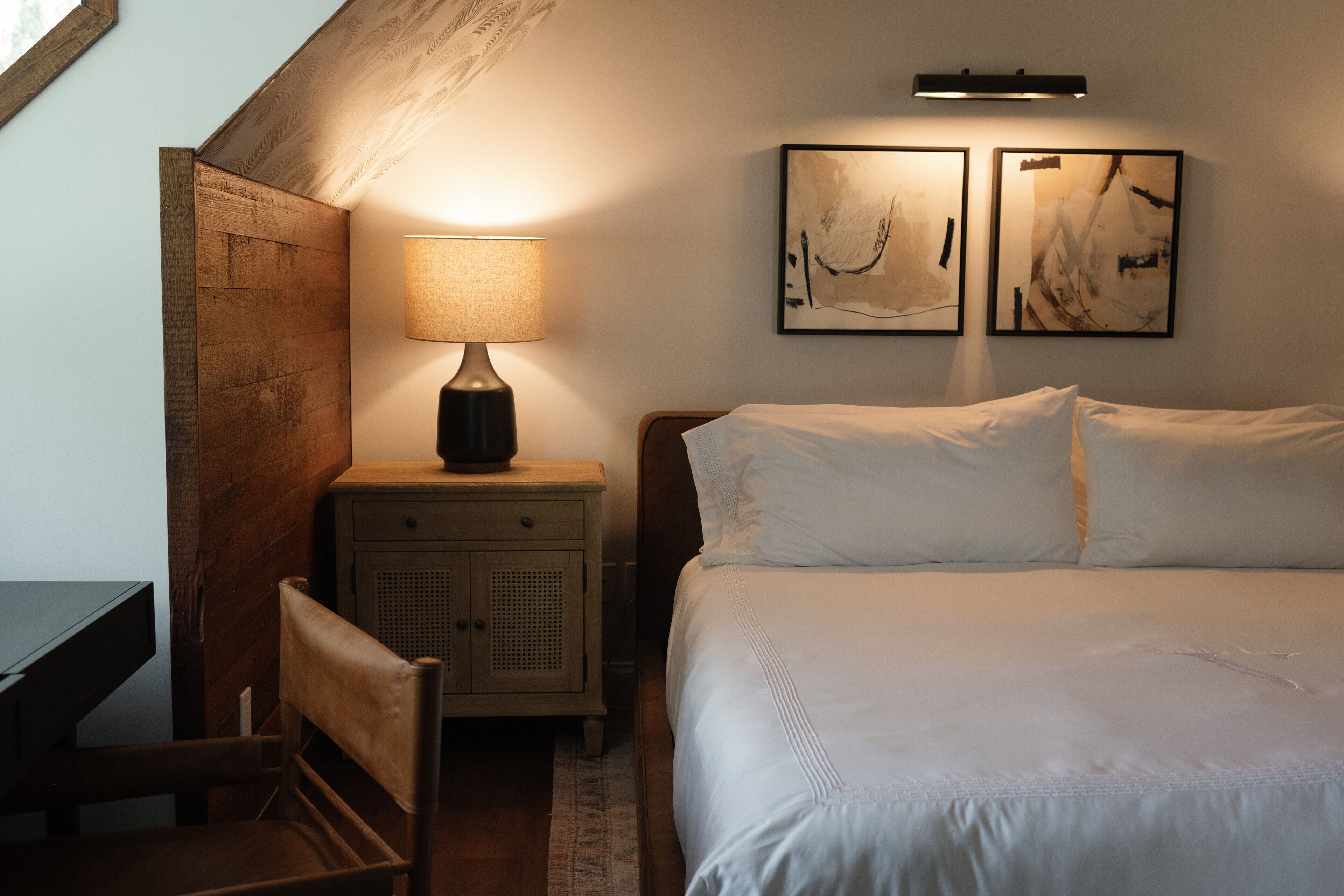 Quarto com cama, mesa de cabeceira com abajur, cadeira e duas pinturas abstratas na parede.