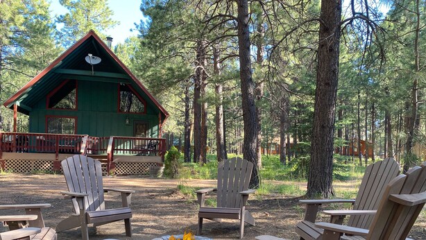 A cabin with a stone fire pit surrounded by Adirondack chairs, set in a forested area. - Bellwood Cabin (Forest Lakes Estates)