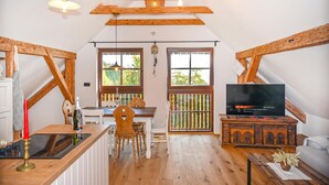 Una accogliente stanza mansardata con soffitto in legno, un'area pranzo con tavolo e sedie, una cucina con fornello e una candela rossa, e un televisore su un mobiletto in legno.