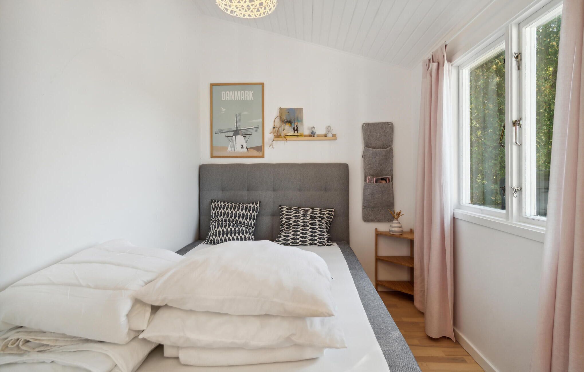 A bedroom with a bed, pillows, a headboard, a shelf with decorative items, a chair, and a window with curtains.