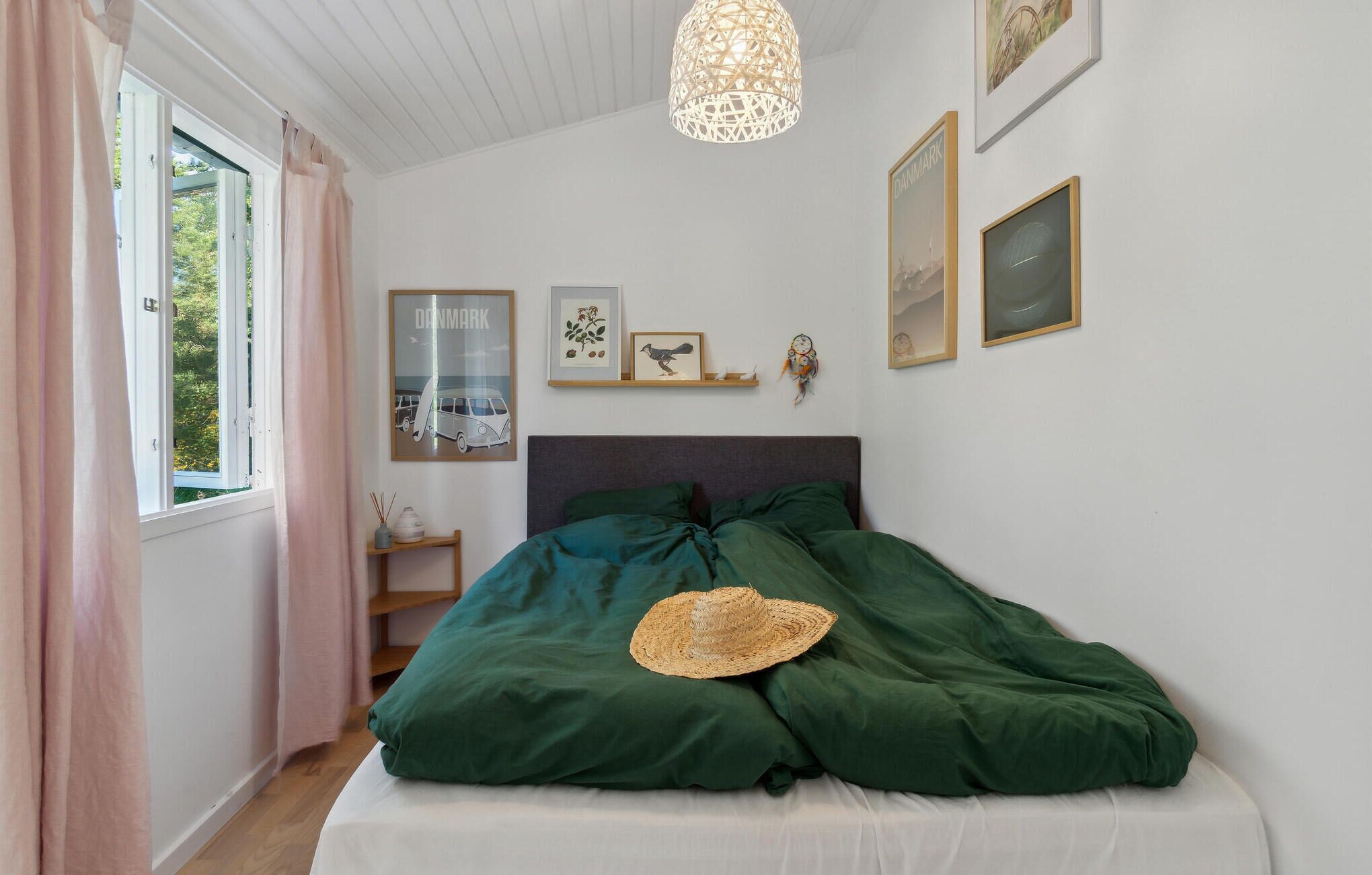 Een slaapkamer met een bed, een raam met gordijnen, een wandplank met foto’s en een hanglamp.