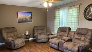 A living room with three reclining chairs, a ceiling fan, a framed picture on the wall, and a window with curtains.