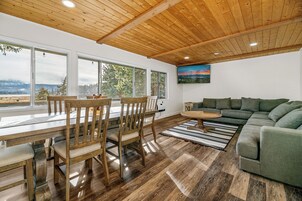 Un salón amplio con techo de madera, ventanas grandes, un sofá verde y un comedor con muebles de madera.