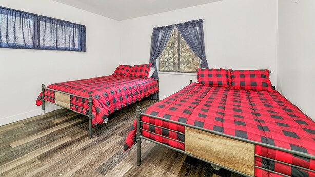 A room with two beds, each with a red and black checkered bedspread, a window with blue curtains, and a wooden floor.