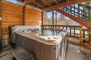 Una terraza de madera con un jacuzzi, barandas de madera y vistas a los árboles circundantes.