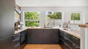 Une cuisine moderne avec des meubles de couleur sombre, un plan de travail en bois et de grandes fenêtres donnant sur un paysage verdoyant.