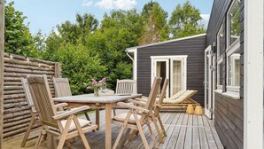 Une terrasse en bois avec une table et des chaises, un petit espace de détente extérieur avec des coussins, et un bâtiment de couleur sombre doté d’une porte et de fenêtres.