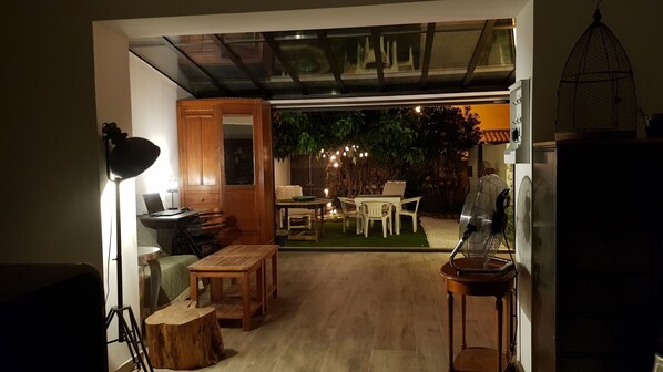 An indoor space with a wooden desk, chairs, a small table, and a lamp. There is a glass door leading to an outdoor area with a table and chairs.
