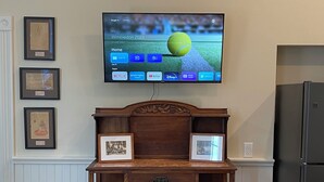 Un salón con televisión instalada en la pared mostrando Wimbledon 2023, un mueble de madera con detalles decorativos, una mesa de centro con un libro y un refrigerador.
