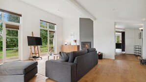 A spacious living room with a grey sofa, a painting on easel, a fireplace, and large windows offering a view of greenery. - Gorgeous home in Jægerspris (Jægerspris)