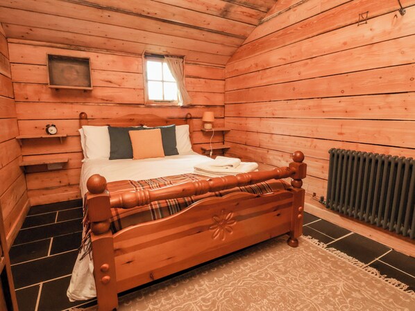 A wooden cabin bedroom with a bed, a nightstand, a window, and a radiator. - Y Caban (Llangollen)