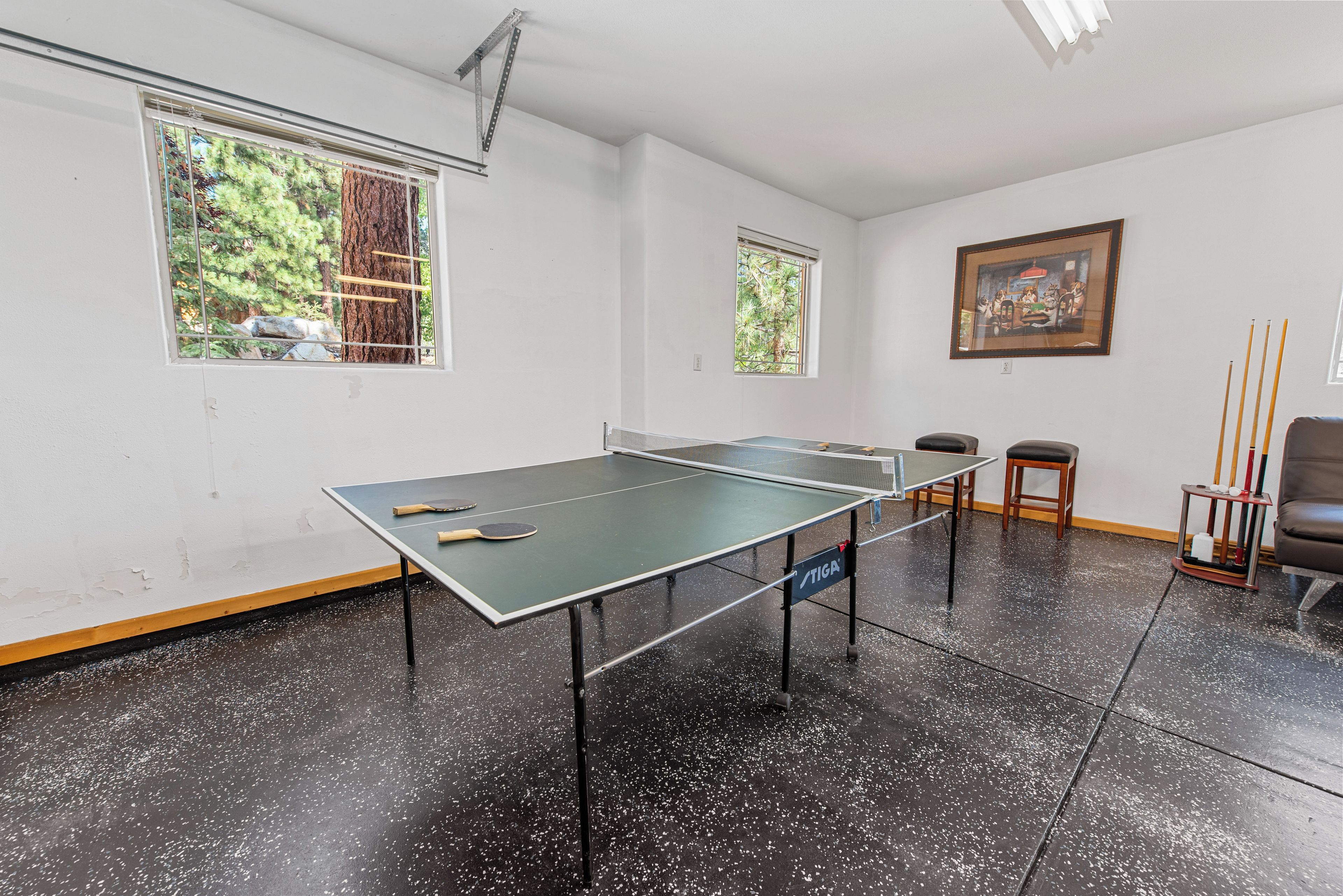 Uma mesa de pingue-pongue em um quarto com duas janelas, um quadro na parede e uma cadeira.