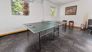 Uma mesa de pingue-pongue em um quarto com duas janelas, um quadro na parede e uma cadeira.