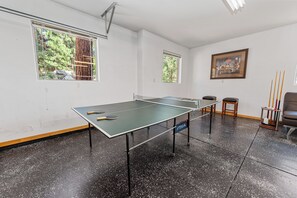 Uma mesa de pingue-pongue em um quarto com duas janelas, um quadro na parede e uma cadeira.